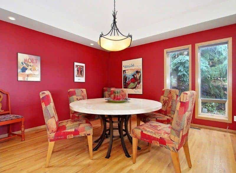 The charming framed vintage posters and artworks area a nice accent for the red walls that is contrasted by a bright white ceiling with recessed lights and a pendant light in the middle hanging over the white round dining table paired with cushioned chairs with colorful patterns.