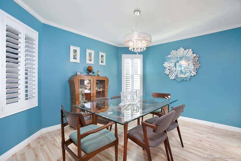 The bright blue walls of this dining room is reminiscent of the blue skies above the beach with the white clouds represented by the white shuttered windows and the sandy beach is the light hardwood flooring that emphasizes the glass-top dining table and its wooden armchairs.