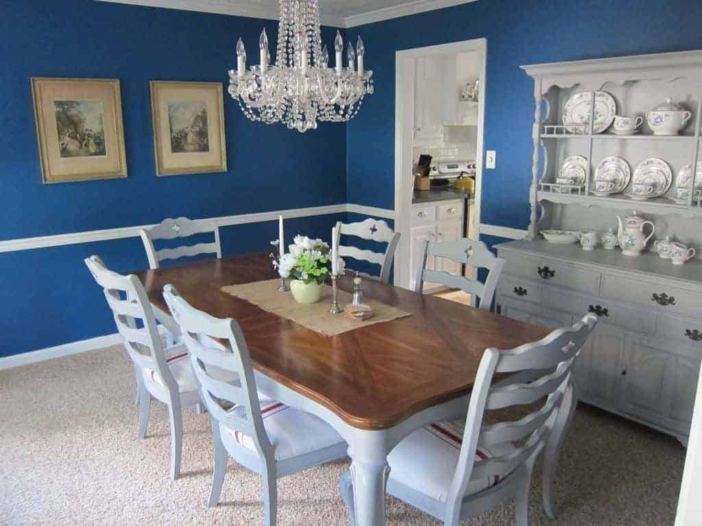 The white wooden chairs surrounding the wooden dining table have a wave design to its back that stands out against the bright blue walls adorned with a couple of classic paintings and a light gray dining room cabinet with a set of chic china dishware on display.