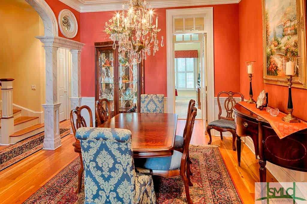 This chic dining room has a majestic crystal chandelier that elevates the elegance of the wooden dining table and its wooden chairs. On each end of this table is a blue patterned cushioned chair that goes well with the colorful patterned area rug and the orange walls.