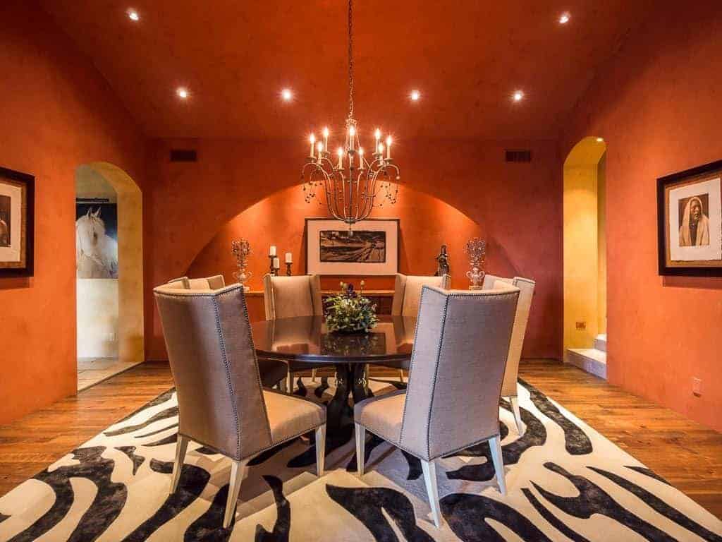The zebra print area rug covering most of the hardwood flooring stands out against the orange walls and ceiling filled with yellow recessed lights and a chandelier that warms up the black wooden dining table and its cushioned chairs of this medium-sized dining room.