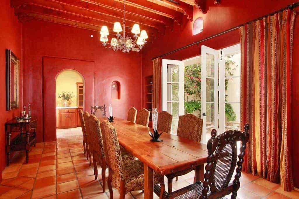 This warm and homey medium-sized dining room has terracotta tiles on its flooring that emphasizes the earthy hues of the long wooden dining table that is paired with wooden chairs that have red patterned cushions complementing the red walls.