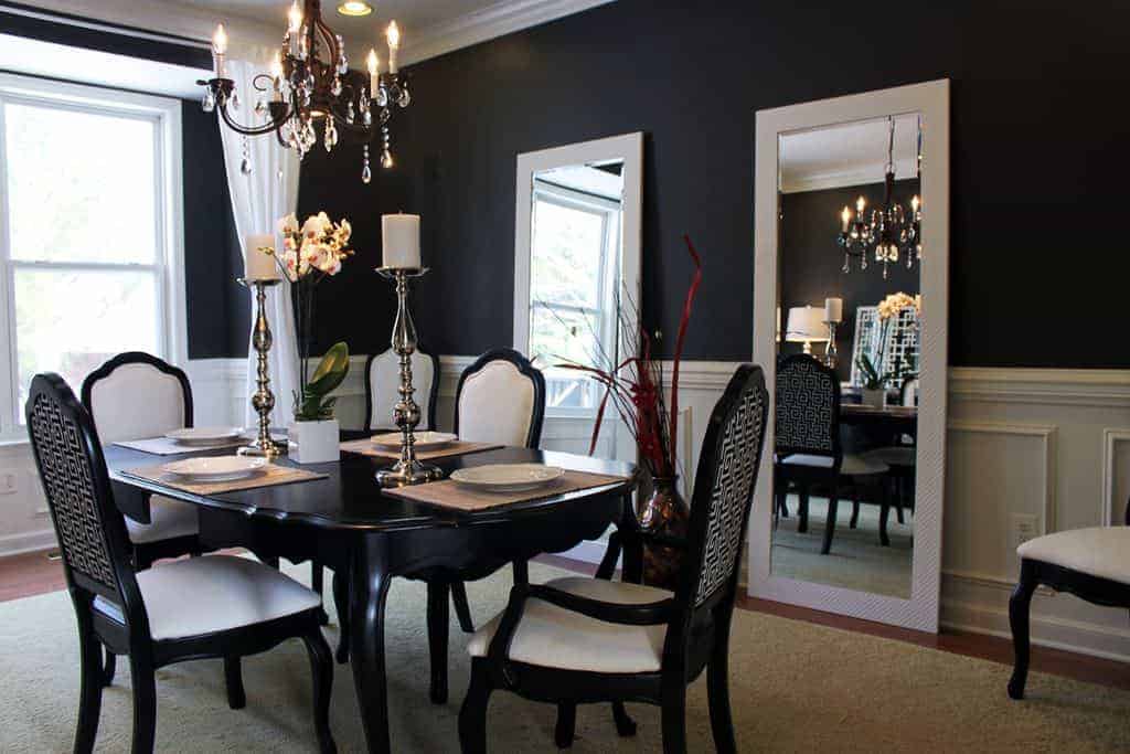 This elegant and formal dining room has black walls contrasted by the white wainscoting and the pair of large rectangular mirrors leaning against the wall by the black wooden dining table and its matching dining chairs with white leather cushions.