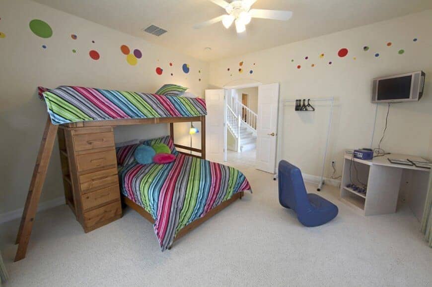 A sleek blue chair faces the custom table and television in this kids bedroom boasting a wooden loft bed dressed in striking multicolored bedding.