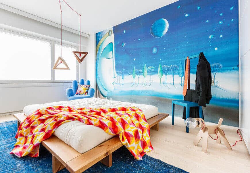A large scale wall mural sets a serene backdrop to the platform bed in this kids bedroom with a blue high back chair and a hand seat lighted by a pair of wooden triangular pendants.