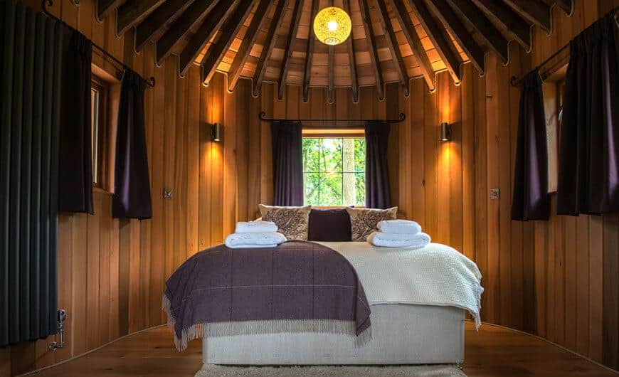 A stunning primary bedroom with rustic walls and ceiling with amazing beams lighted by warm lighting. The room has a gorgeous bed setup.