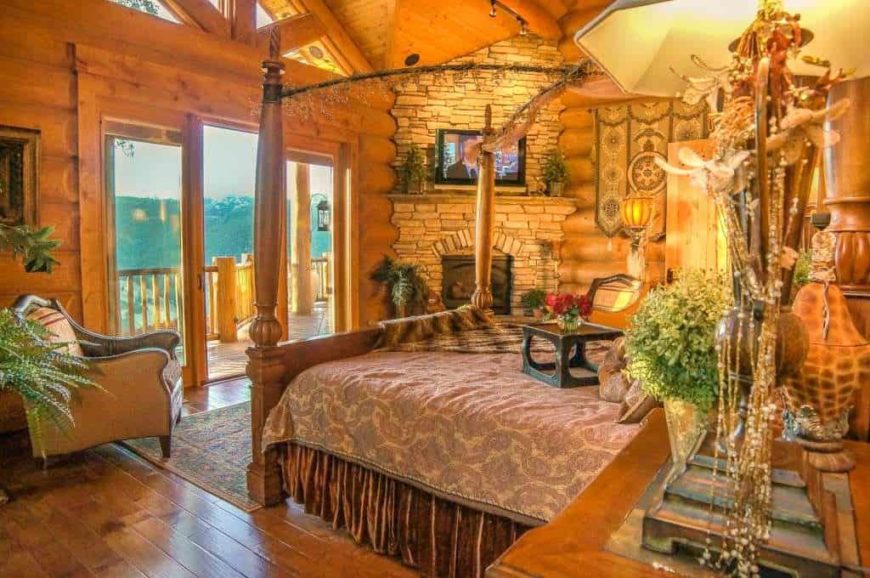 Large primary bedroom with rustic walls, ceiling and flooring. It has an elegant bed setup and a stone fireplace with a TV on top. The warm lighting looks so perfect together with the bedroom's rustic style.