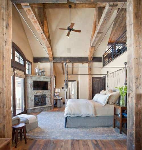 Rustic primary bedroom featuring a tall ceiling with large rustic beams. The room also features hardwood flooring topped by a large gray rug along with a fireplace with a widescreen TV on top.