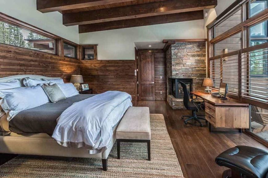 Large rustic primary bedroom with hardwood flooring and wooden walls, along with a wooden ceiling with rustic beams. The room has a large comfy bed and an office area featuring a rustic office desk.