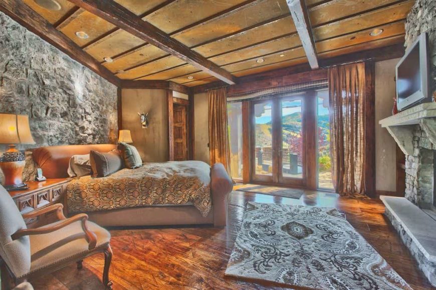 Large rustic primary bedroom with a stone wall, hardwood flooring and a rustic ceiling. The room has a large cozy bed lighted by two table lamps and a large stone fireplace.
