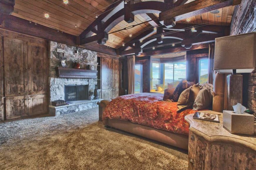 Large rustic primary bedroom featuring a stunning wooden ceiling with large beams along with carpeted flooring. The room offers rustic furniture along with a large fireplace.