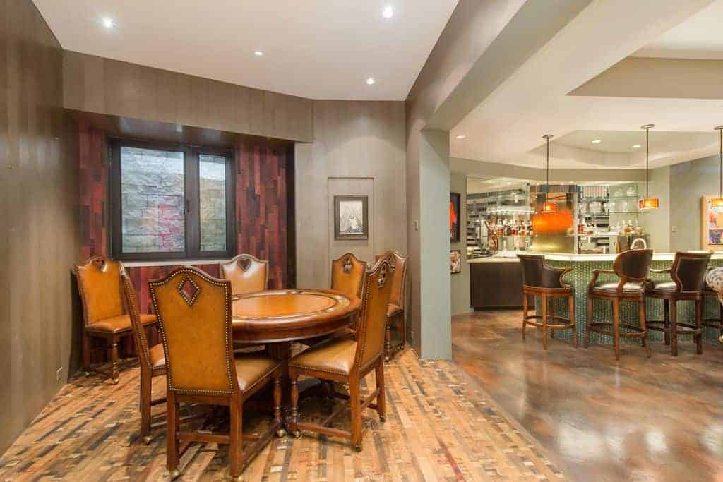 This dining area is inside an irregular shaped alcove surrounded by dark gray walls, white ceiling with recessed lights and a unique hardwood flooring that somehow pairs with the redwood wall that supports the window looking over the brown leather dining chairs.