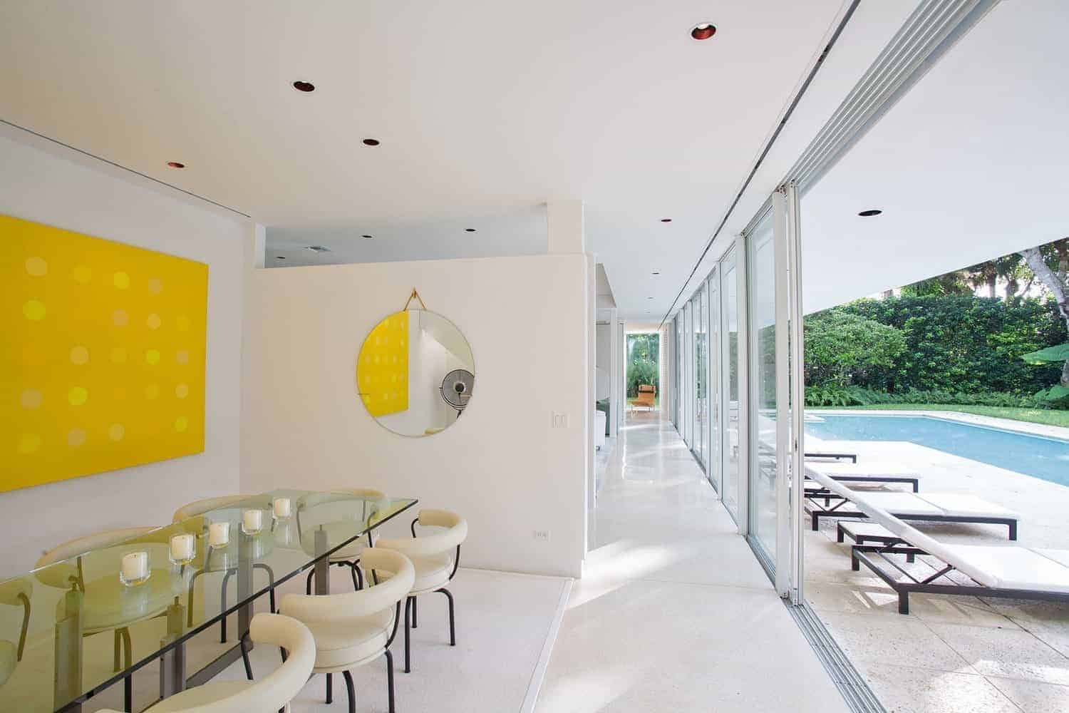 The glass-top rectangular dining table is a perfect match for the sliding glass doors that open to a poolside scenery. This gives a sense that the dining room is bigger that it seems. The addition of the yellow abstract painting complements the beige modern dining chairs.