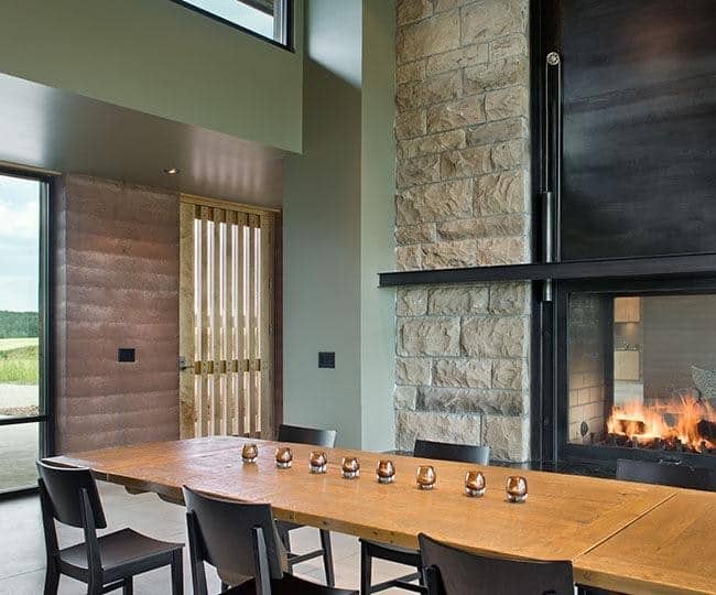 The long rectangular wooden dining table is paired with black wooden chairs that lend with the frame of the fireplace that is fixed against a large pillar of beige stones that stands out beside the light green hue of the walls but matches with the beige flooring tiles.