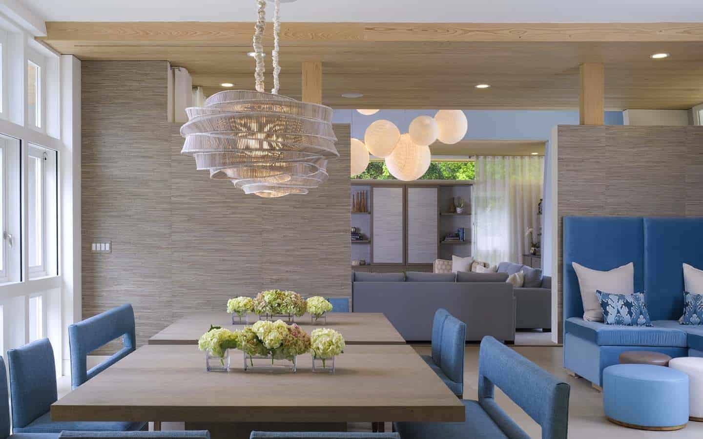 This airy and bright kitchen has a bare wooden ceiling paired with brown wooden walls that match the square dining tables topped with a peculiar pendant light with a rustic hood. These are contrasted by the blue velvet cushions of the chairs.