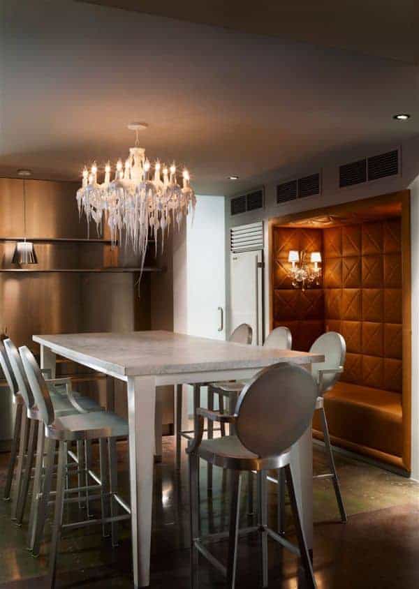 The stainless steel modern stools are paired with a tall rectangular table that has a white marble top contrasting the dark hardwood flooring but matches with the white chandelier that has a peculiar design that make it seem like a melting cake frosting.