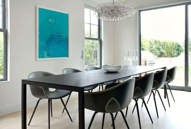 This dining room has a simple dining set that is comprised of a long modern black rectangular table and modern black dining chairs. This stands out against the beige flooring and white walls that is adorned with a blue painting flanked by bright windows.