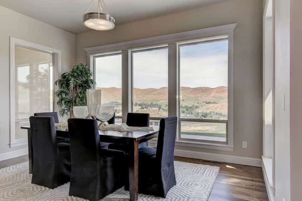 The dining chairs of this medium-sized dining room has black velvet slip covers that stand out against the light gray patterned area rug and the beige walls that are dominated by tall windows that showcase a nice mountain view.