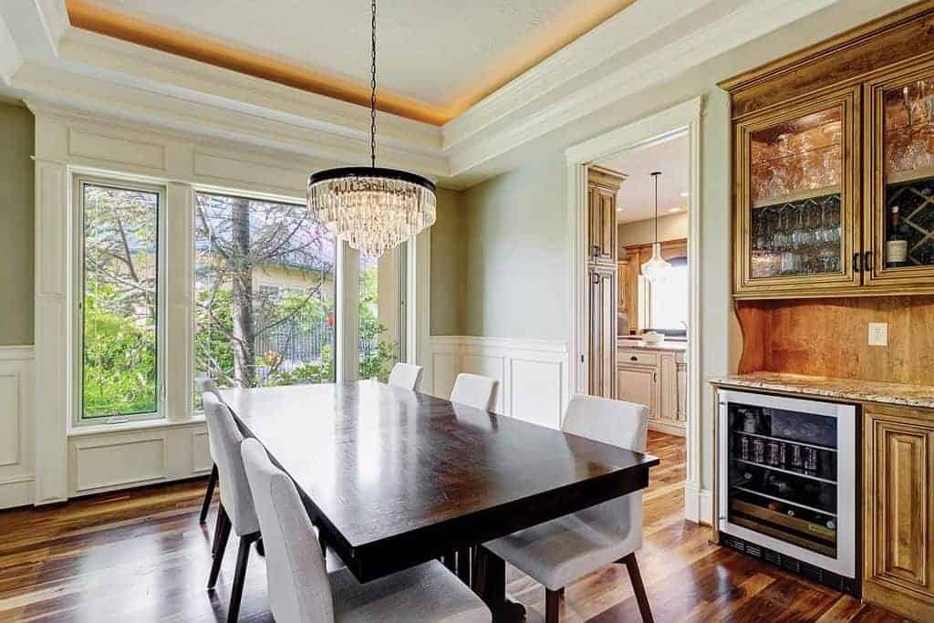 The dark wooden rectangular dining table is matched in elegance by the crystal pendant light hanging from the white tray ceiling. This matches with the white wooden finishing of the walls brightened by a large glass window.