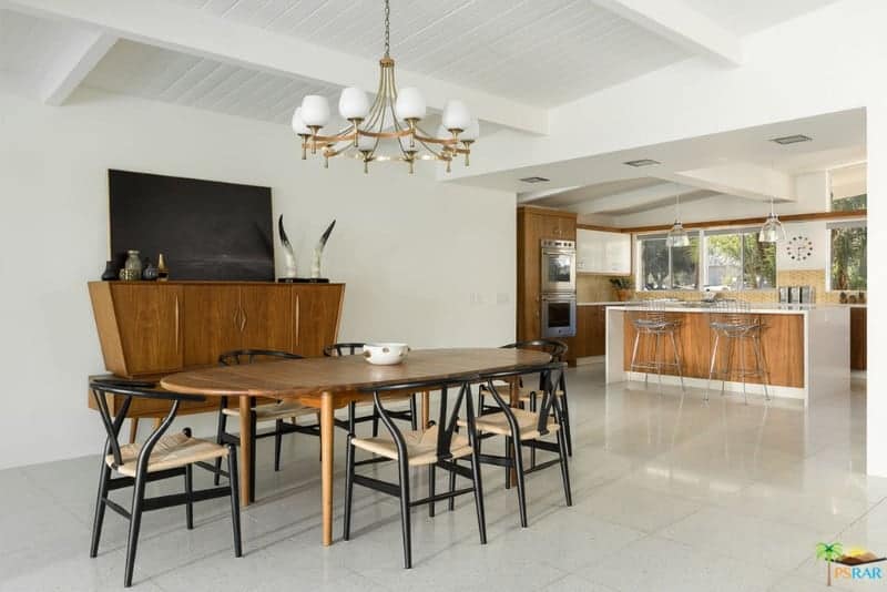 The bright white shiplap ceiling has exposed beams that matches the bulb hoods of the brass chandelier. This is a complement to the wooden dining table and the console table that has built-in cabinets. This is contrasted by the black wishbone chairs that have rustic woven wicker seats.