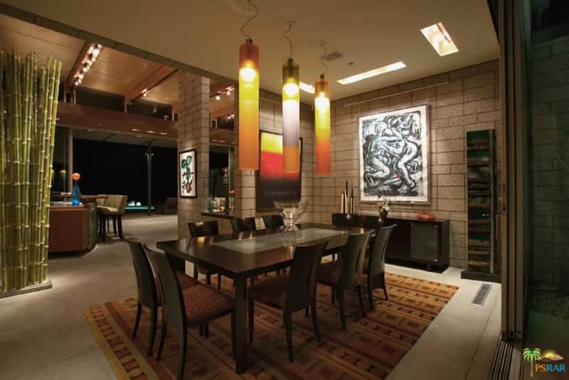 The highlight of this medium-sized dining room is the trio of tube-like pendant lights that have a colorful orange hue to them that stands out against the dark wooden dining set that matches with the console table, area rug and even the stone walls.