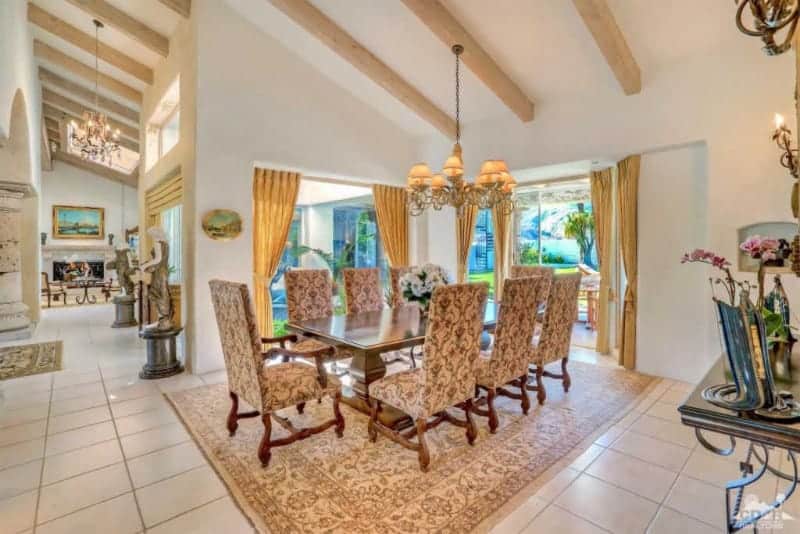 The white shed ceiling has exposed wooden beams that support an intricate chandelier that matches the chic quality of the dining chairs and their patterned cushions as well as the beige patterned area rug over the white tiles of the flooring.
