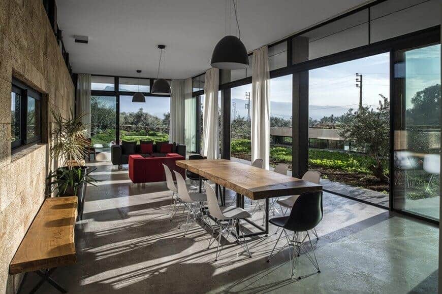 This medium-sized dining area shares its industrial-style gray flooring and white ceiling with the living room area. These two area are bathed in sunlight from the tall glass windows. The thick wooden dining table matches with the wooden bench against the brown stone wall.