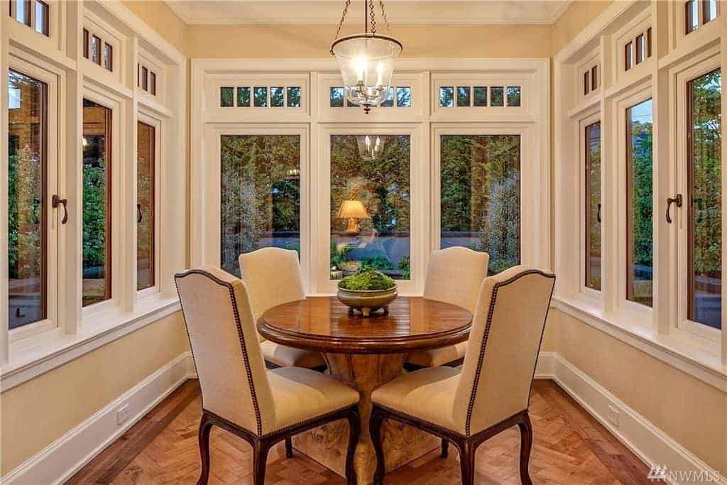 The beige cushions of the dining chairs are a perfect match for the beige walls. These parts are barely seen due the dominance of the white tall windows surrounding the medium-sized dining room. The hardwood flooring that has a herring bone pattern matches the wooden round table.
