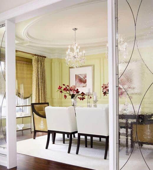 This dining room is separated from the rest of the house by frosted glass walls with white frames that match the white cushioned dining chairs, area rug on the dark hardwood flooring and the white ceiling with an elliptical design that supports a white chandelier in the middle.