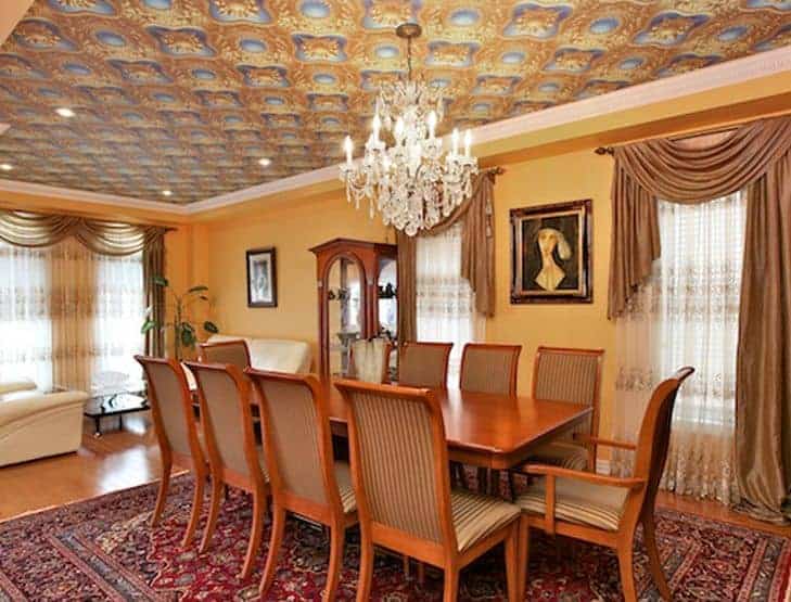 The warm yellow walls that are accented with classic paintings match with the yellow details of the complex and peculiar design of the ceiling. It has a majestic white crystal chandelier that brightens up the wooden dining set and colorful patterned area rug.