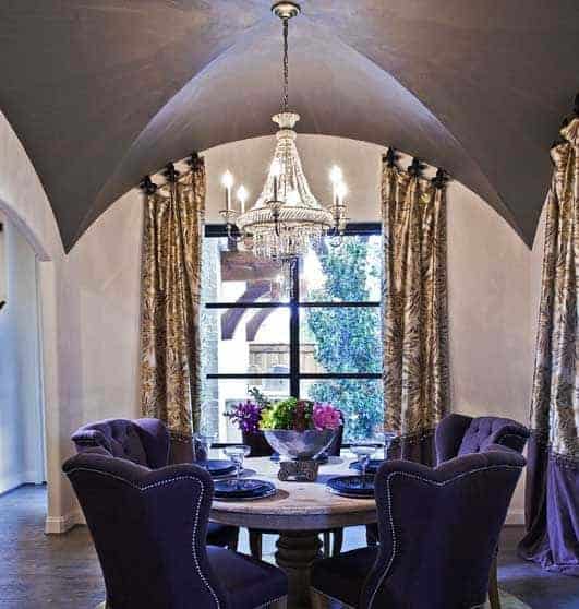 The circular wooden table is encircled with purple velvet cushioned chairs that match with the curtains that has a purple design on its edges complementing the dark hardwood flooring illuminated by the majestic chandelier from the groin arch ceiling.