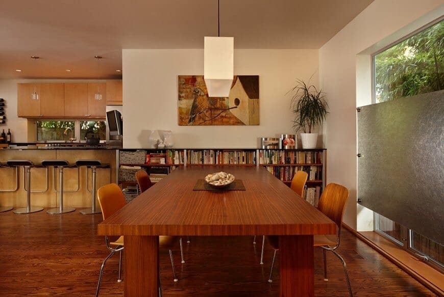 The beige wall above the beautiful bookshelf is adorned with a painting of a bird and its birdhouse that has various shades of brown. This painting sets up the brown theme of the dining area that has a brown wooden rectangular table and brown modern chairs over a hardwood flooring.
