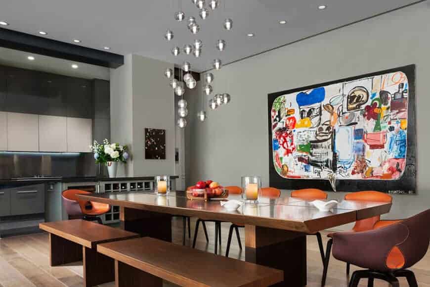 This informal dining room has a friendly and quirky demeanor with its colorful painting on the light gray walls and small silver spherical pendant lights hanging from the gray ceiling. This colorful scheme is matched by the orange modern dining chairs of the wooden table and its wooden benches.