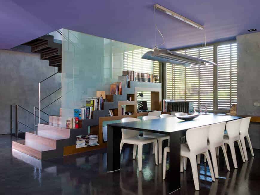 This modern dining area by the stairs has white modern chairs that stand out against the black modern dining table and black industrial-style flooring that matches well with the industrial-style fluorescent light hanging from the ceiling.
