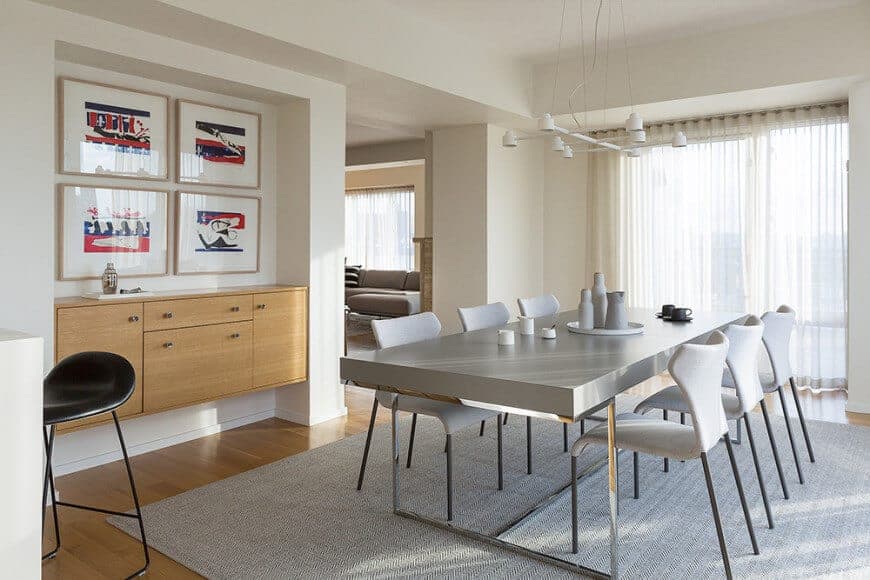 This gorgeous dining room is a wonderful fusion of traditional and modern elements. It has a modern white chandelier hanging over the modern dining set with stainless steel legs complementing the gray area rug over the hardwood flooring that matches the floating cabinet embedded into the white wall on the side.