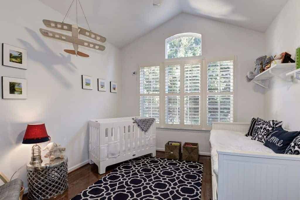 White nursery contrasted with black patterned rug and pillows that lay on the beadboard sofa. It has a sleek crib and stylish round table topped with a red lampshade.