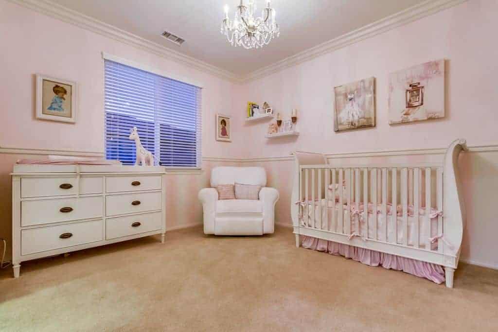 Charming wall arts hang above the skirted crib in this light pink nursery with white drawer chest and armchair lighted by a crystal chandelier.
