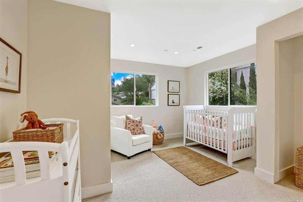 Taupe nursery with carpet flooring and glazed windows overlooking the outdoor greenery. It has white furnishings and a jute runner that complements the rattan baskets.