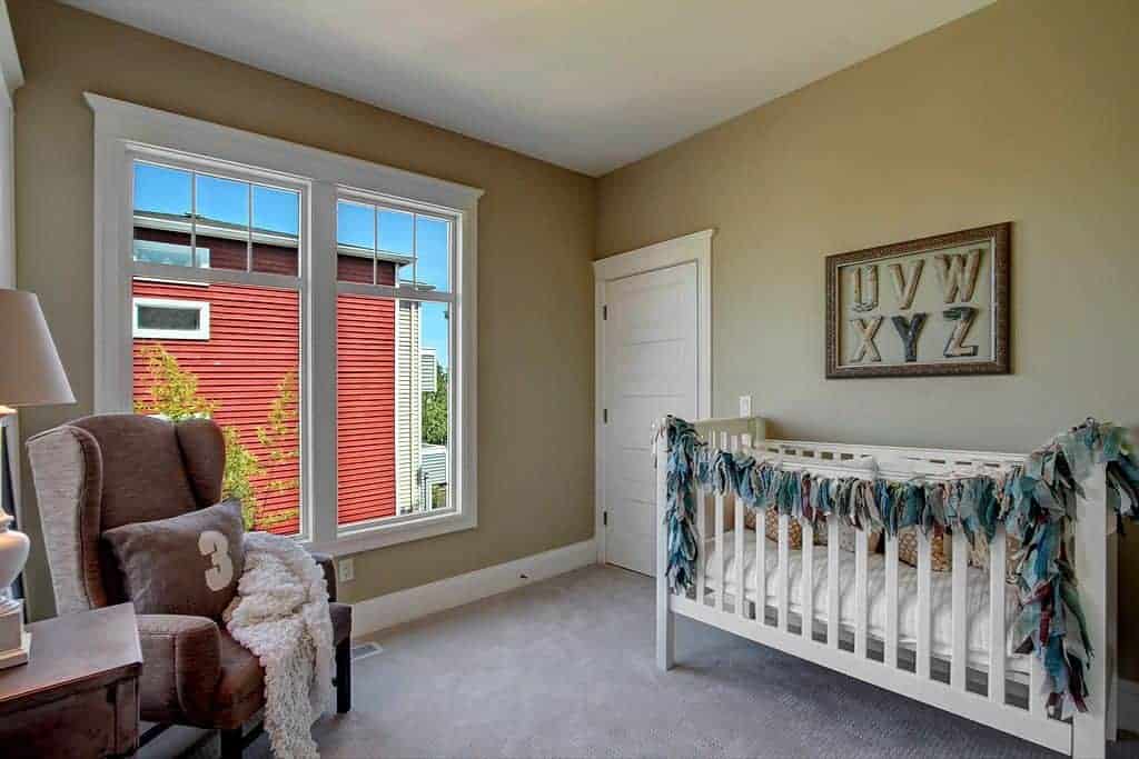 This nursery features a brown wingback armchair and white crib accented with an alphabet artwork. It has beige walls and framed windows inviting natural light in.