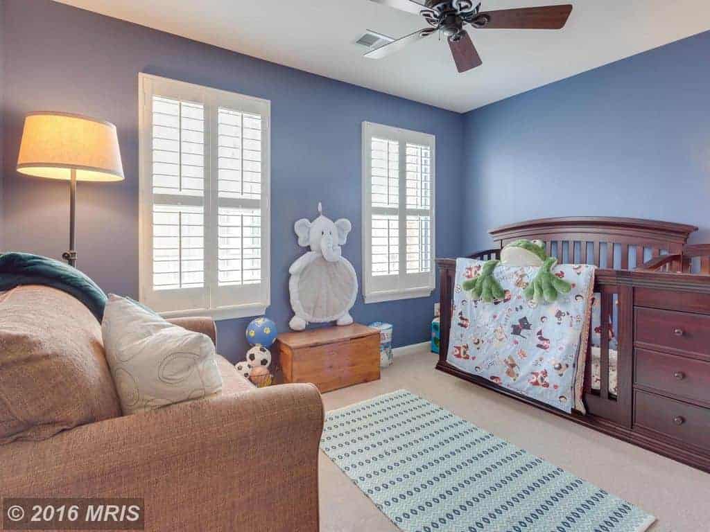 Blue nursery with a comfy armchair and wooden crib fitted with built-in drawers. It includes a ceiling fan and patterned runner that lays on the gray carpet flooring.