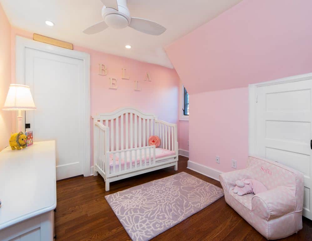 A traditional table lamp sits on the white cabinet in this pink nursery showcasing a charming crib and dotted armchair with a purple floral rug in the middle.