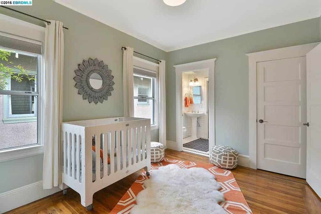 Light and airy nursery designed with layered rugs and a gorgeous floral mirror mounted above the white crib. It has hardwood flooring and picture windows covered in white curtains.
