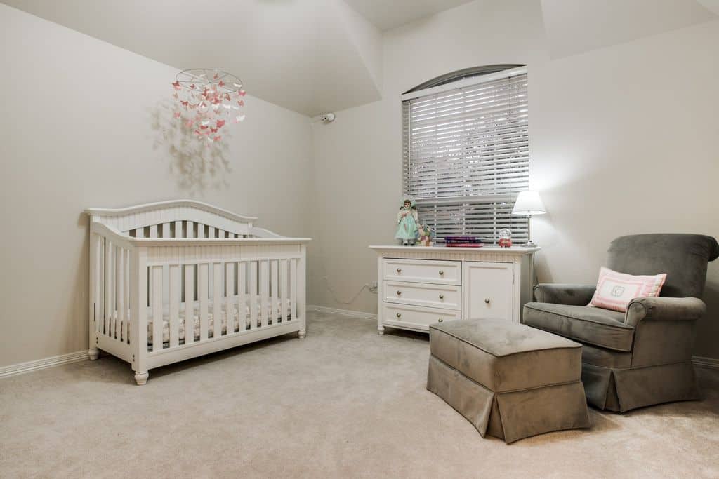 A gray skirted ottoman matches the velvet armchair in this minimalist nursery offering a white cabinet and crib paired with a pink butterfly mobile.