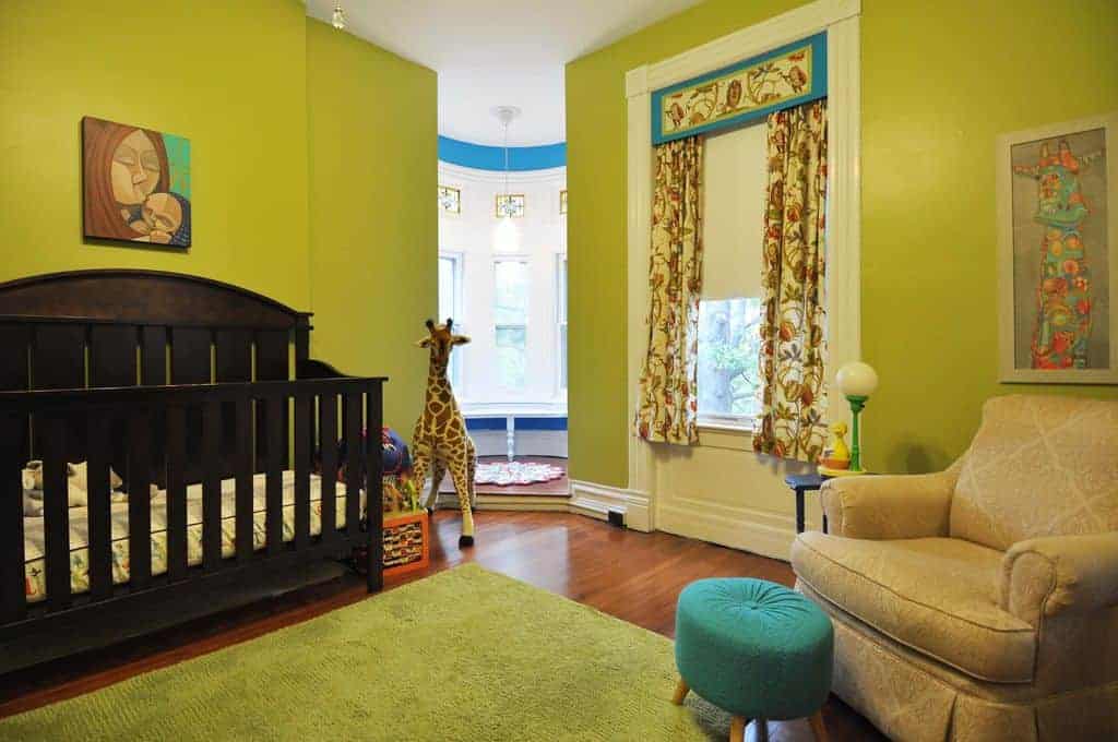A vibrant green nursery boasts a skirted armchair paired with a round ottoman along with a dark wood crib accented with a mother and child painting.