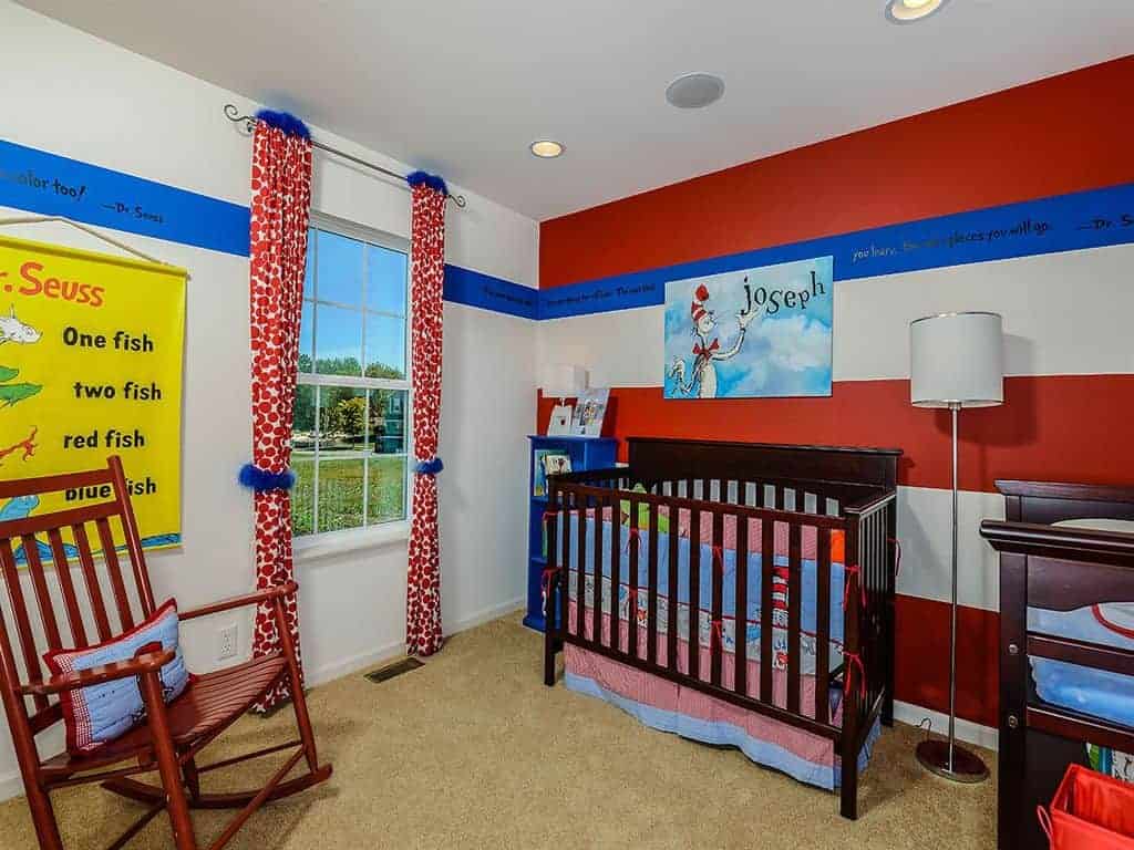 This nursery features a wooden rocking chair and skirted crib lighted by a chrome floor lamp. It has carpet flooring and white framed windows dressed in red dotted draperies with blue fur accents.