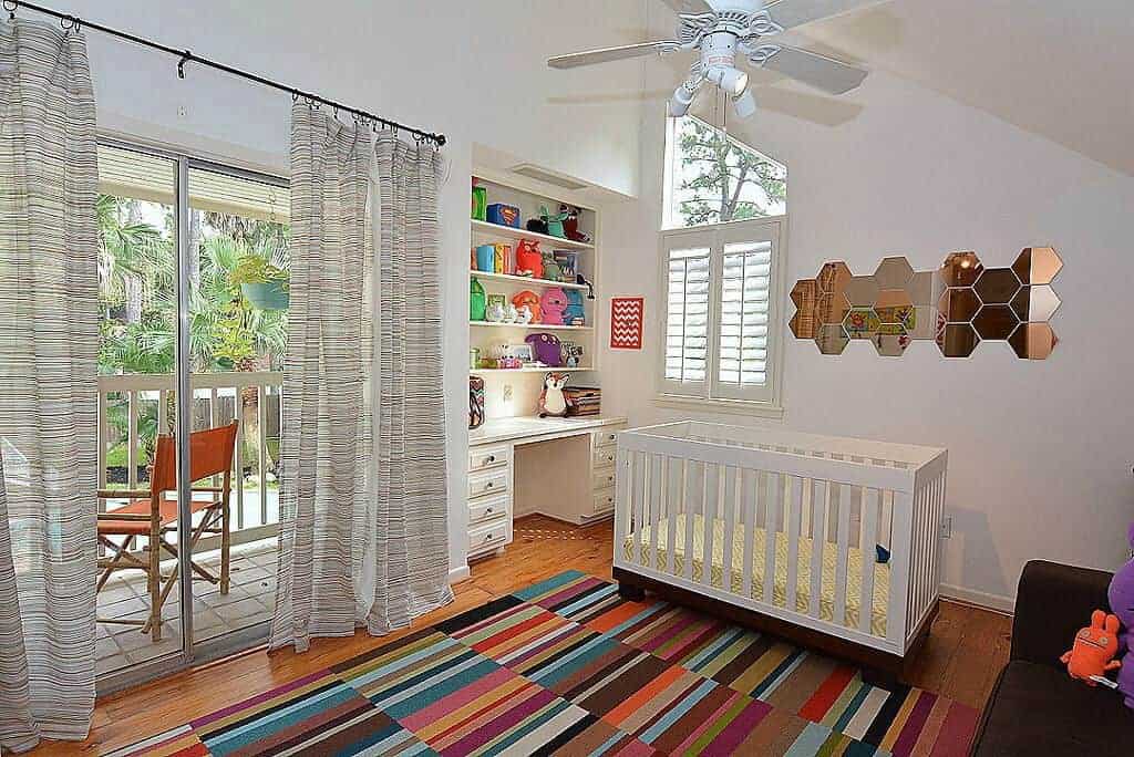 A colorful striped rug adds a striking accent in this white nursery with a desk and stylish honeycomb mirror mounted above the sleek crib. It has built-in shelving and glass slider that leads out to the balcony.