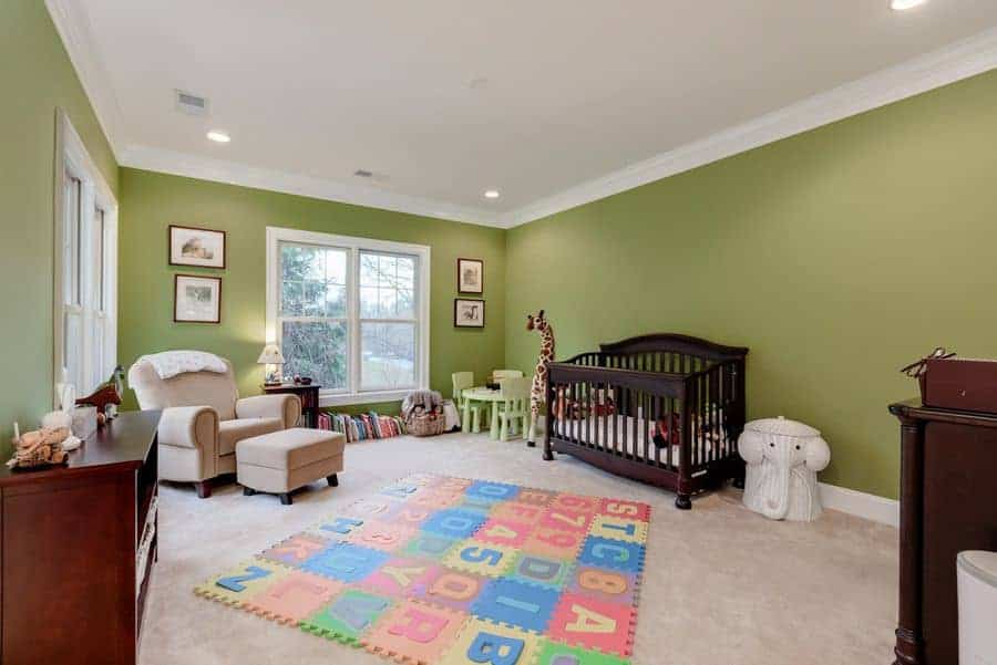 A large nursery with green walls and gray carpet flooring topped by an alphabet puzzle mat. It has smooth armchair with matching ottoman and a dark wood crib next to the small table and chairs.