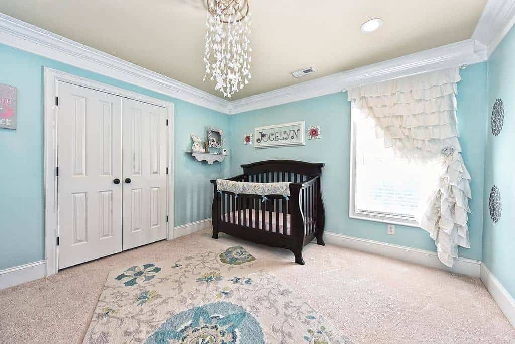 A white double door opens to this aqua nursery showcasing a charming floral rug and dark wood crib with a framed baby's name on top.