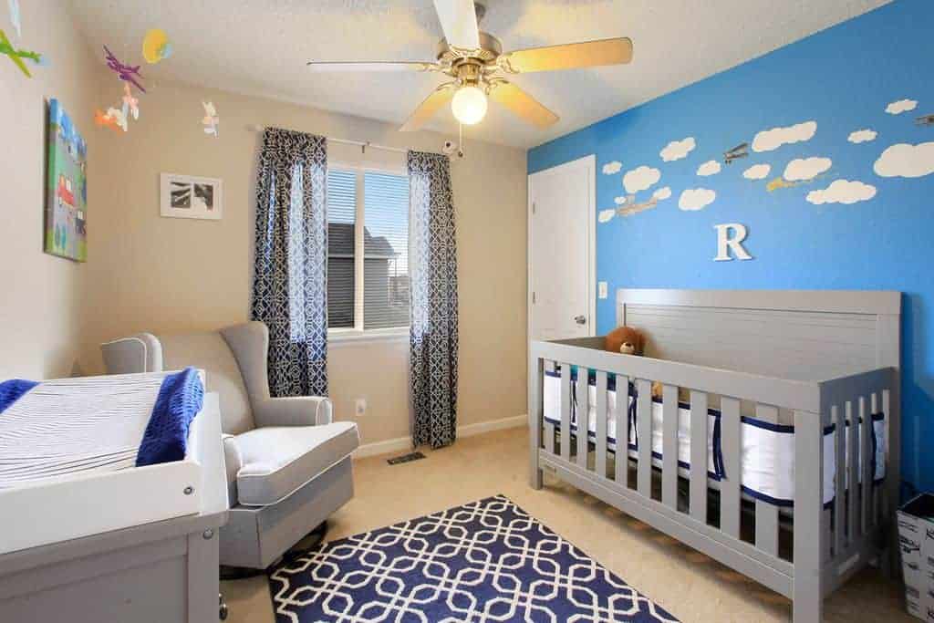 Patterned draperies complement the deep blue area rug in this beige nursery with wingback armchair and gray crib placed against the cloud accent wall.