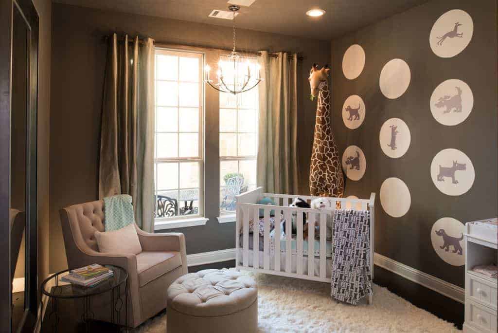 Gray nursery decorated with a candle chandelier and a large giraffe plush toy that sits behind the white crib on a shaggy area rug. It has full height mirror and a tufted armchair accompanied by a matching round ottoman and glass top side table.
